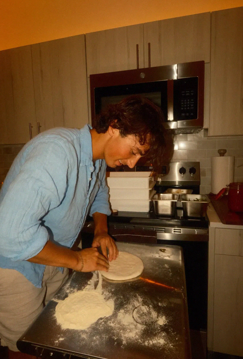 Malachi smiling as he presses out pizza dough on a floured steel table
